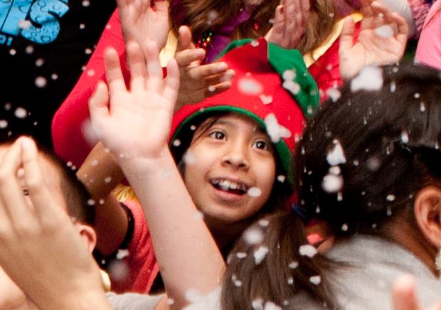 A smiling child in an elf hat reaching up joyfully as snow falls around them