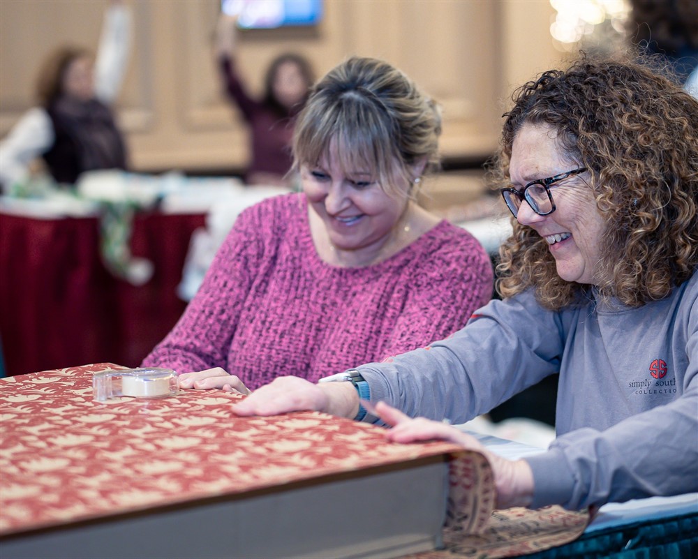 Two volunteers laughing together while wrapping a large gift in red patterned paper at an Operation North Pole gift wrapping event
