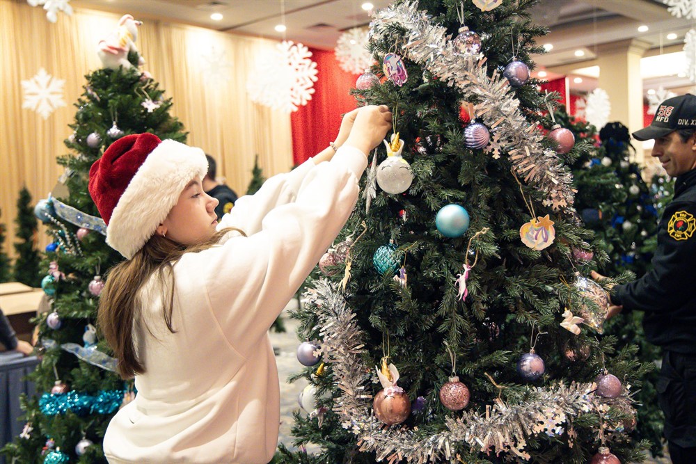 A young volunteer in a Santa hat hanging ornaments on a decorated Christmas tree at Operation North Pole