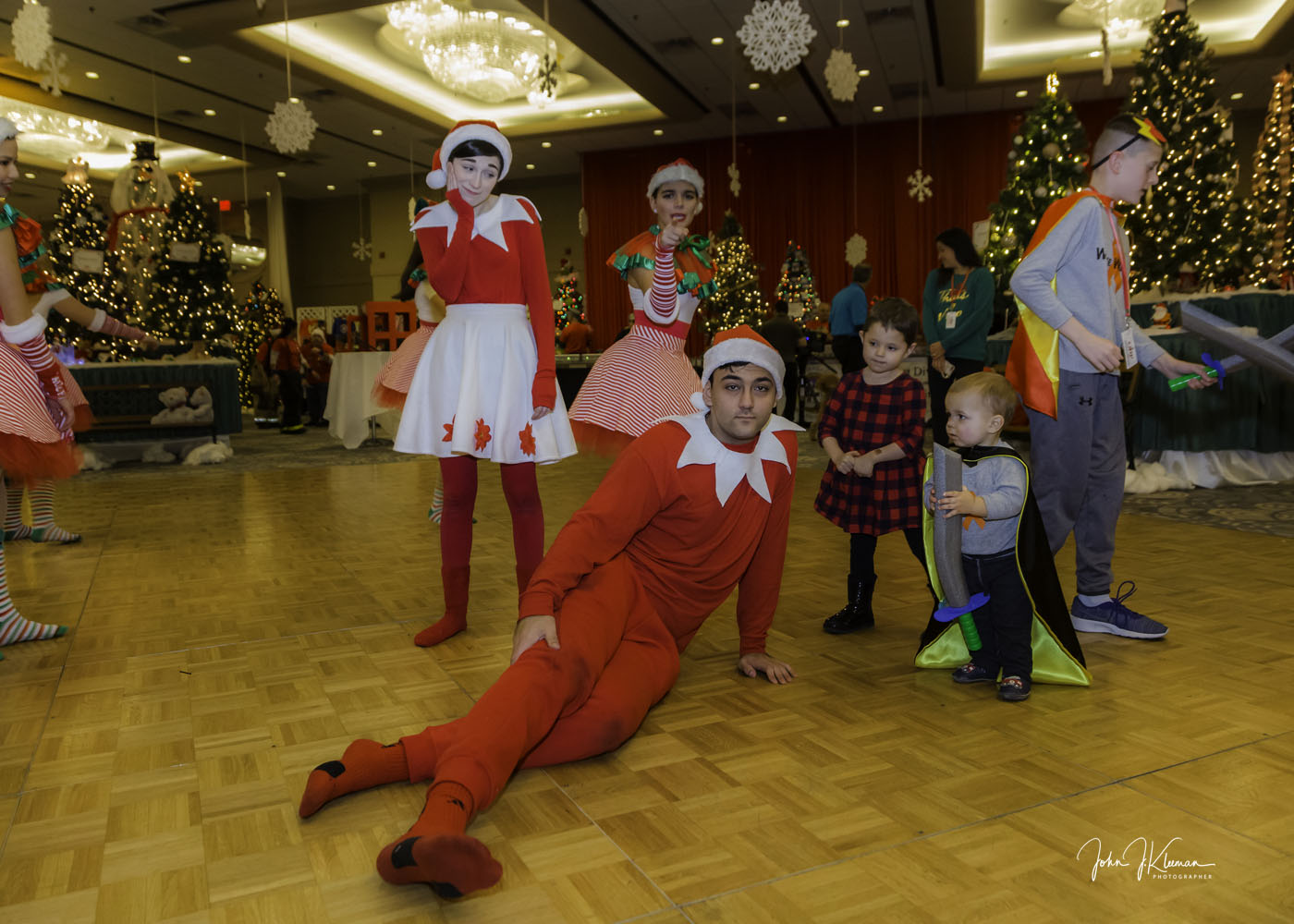 Elf performers dancing with children in a ballroom of Christmas trees