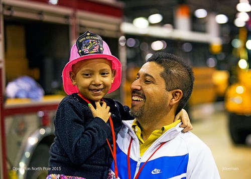 A smiling father holding his daughter who is wearing a pink North Pole Fire Academy helmet